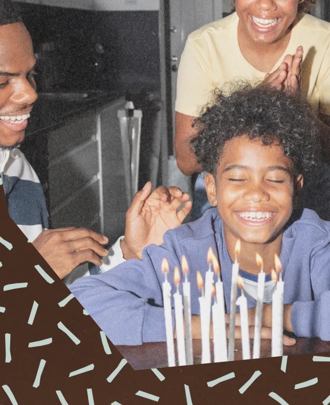 A boy laughing in front of a candlelit birthday cake with his parents behind him, against a sprinkled background.
