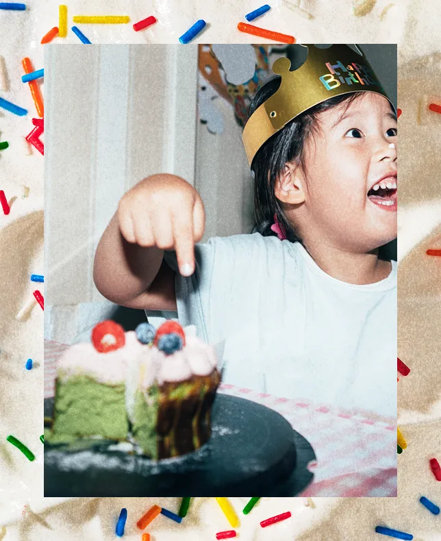 A young girl in a birthday crown digging into a small cake, against a colorful sprinkles background.
